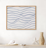Abstract wall print with smooth, wavy blue lines on a pale background, framed in light wood, displayed above a neutral-toned surface with coastal decor accents.