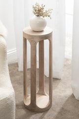 Tall, slender wooden drink table with curved rectangular cutouts in the legs and a round top holding a white ceramic vase with dried branches, against a white curtain backdrop.