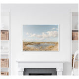 Framed canvas print of a serene marshland with tall golden grasses, calm blue water, and a partly cloudy sky, displayed above a white mantel.