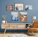 Five framed artworks on a blue wall above a light wood sideboard and leather chair, featuring underwater coral, abstract colors, ocean wave, underwater plants, and coastal path scenes.