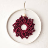 Burgundy macrame snowflake ornament with fringed edges and a small wooden bead, suspended by a natural twine loop on a white ceramic plate.