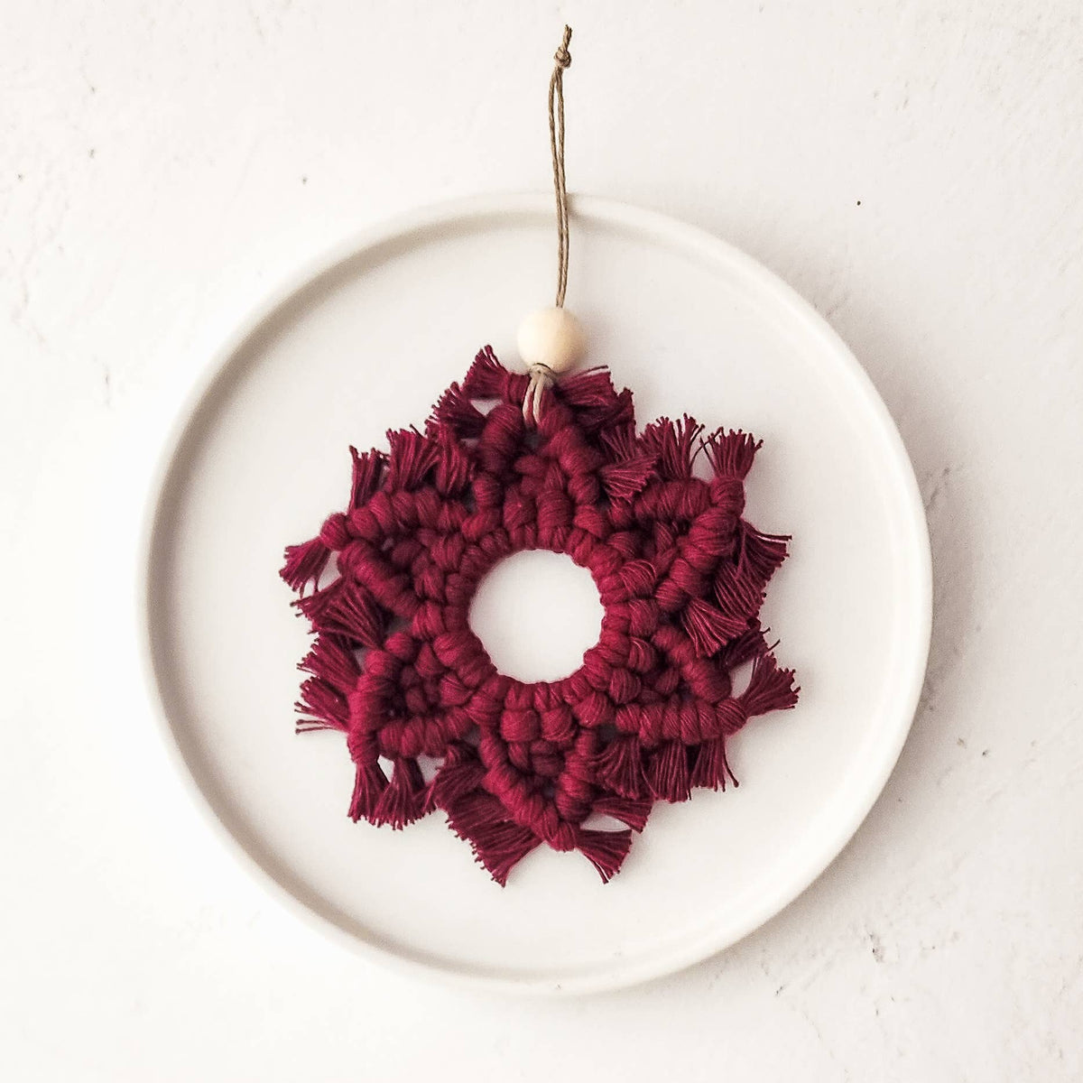 Burgundy macrame snowflake ornament with fringed edges and a small wooden bead, suspended by a natural twine loop on a white ceramic plate.