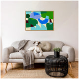 Abstract geometric painting with blue, green, and brown shapes in a natural wood frame above a beige sofa with a gray cushion, blanket, and a black wireframe coffee table with a small plant.