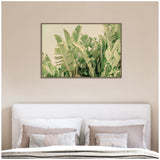 Framed canvas print of green tropical banana leaves against a light sky, hung above a bed with beige and gray pillows and soft linens.