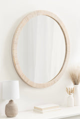 Oval wall mirror with textured light beige frame hanging above a white shelf holding a small lamp, stacked books, and two vases with dried grasses.