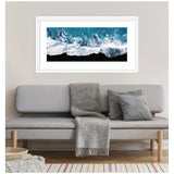 Framed aerial art panel showing turbulent ocean waves crashing onto a black sand beach with white foam and deep blue water above a light gray sofa with gray cushions and a throw blanket.