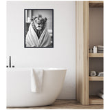 Black and white framed canvas of a lion wrapped in a towel hanging on a white textured wall above a white bathtub with a towel draped over its edge in a modern bathroom.