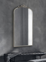 Tall arched wall mirror with ornate top detailing in muted silver, hanging above a black tufted bench with a folded black and white patterned throw.