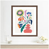 Framed colorful abstract floral print featuring pink vase and various stylized flowers in green, blue, orange, and purple on off-white background, displayed above white console with glass cup and small flower arrangement.