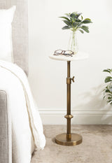 Small round white marble table with adjustable brass pedestal base, holding a clear vase with green leaves and glasses, positioned next to a white bed.