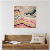 Abstract square canvas with flowing, layered bands in muted earth tones, blues, pinks, and creams, framed in light wood, hanging above a rustic wooden bench with a 'welcome home' pillow.