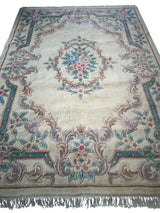Rectangular hand-tufted wool rug with a central floral medallion and ornamental border in soft pastel blues, pinks, and purples on a beige background with fringe ends.