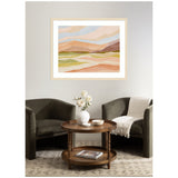 Abstract landscape art with layered pastel hues of peach, beige, green, and lavender, framed and hung above a seating area with dark gray chairs and a wooden round table.