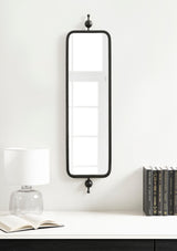 Tall rectangular wall mirror with rounded black metal frame and decorative finials on top and bottom, mounted above a white surface with lamp and books.