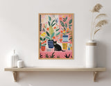Colorful framed artwork featuring a whimsical black cat surrounded by various patterned houseplants on a pink and peach background, displayed above a minimalist wood shelf with white vases and pampas grass.