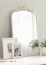 White arched mirror with ornate carved detail atop, placed on a white surface alongside two white photo frames and a green leafy plant in a white pot.