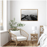 Coastal beach boardwalk in black and white, framed in wood, hanging above a wicker chair and wooden nightstand with white lamp in a bright room with wooden flooring.