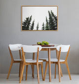 Framed canvas of dark green evergreen tree tops against white background hangs on gray wall above a round wood dining table surrounded by four wooden chairs with white seats and backs.