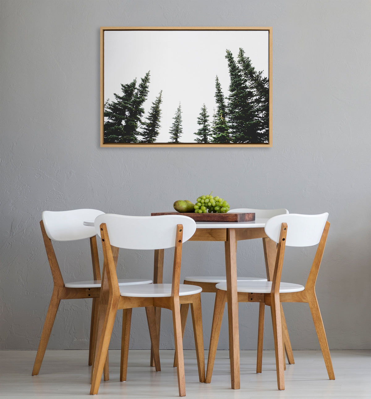 Framed canvas of dark green evergreen tree tops against white background hangs on gray wall above a round wood dining table surrounded by four wooden chairs with white seats and backs.