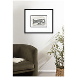 Black-framed grayscale print of Venice's Rialto Bridge with gondola, hanging on a white wall above a dark green armchair and potted olive tree.