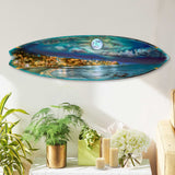 Oval surfboard wall art depicting a luminous full moon over a vibrant coastal town with clouds and ocean waves, mounted on a white wall above plants and candles.