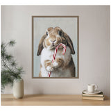 Framed portrait of a brown and white rabbit holding a red and white candy cane against a pale background, hanging on a beige wall above a wooden surface with a vase and stacked books.