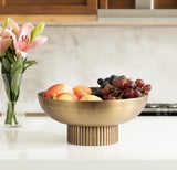 Brushed metal bowl with a ribbed cylindrical base holding peaches and red and black grapes on a white kitchen countertop with pink lilies in the background