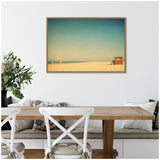 Framed canvas print of a minimalist beach scene with a small lifeguard tower on sandy shore under a clear gradient blue sky, hanging above a wooden dining table.