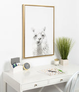 Softly detailed gray and white alpaca portrait centered in a simple light wood frame, hanging above a white desk with minimal office accessories and a green plant.