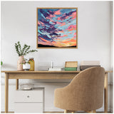 Square canvas painting of a colorful sunset with pink, orange, blue, and purple clouds above a dark horizon, framed in natural wood and hung above a wooden desk.