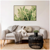 Light beige sofa with a small dog sitting among a gray throw and a round beige cushion, black rattan-style coffee table with a small green plant, large framed green leafy plant art above.
