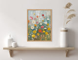 Framed boho-style wildflower garden print with colorful flowers in red, yellow, pink, and white against a soft blue background hanging above a wooden shelf with minimalistic vases and pampas grass.