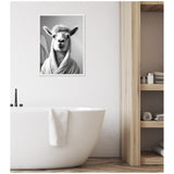 Black and white framed canvas of a llama wearing a bathrobe, hanging on a white wall above a modern white bathtub with a towel draped over it, next to wooden open shelves.