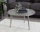 Round coffee table with an uneven, textured gray metal top and three slender metal legs, placed on a white rug in front of a dark gray sofa.
