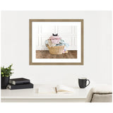 Framed photograph of a wicker laundry basket filled with pastel-colored clothes and a black cat peeking over the top, against white paneled wall and wooden floor.