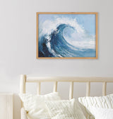 Framed abstract painting of a large ocean wave in shades of blue and white, hanging on a white wall above a light wood bed and cream pillows.