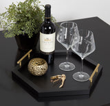 Black hexagon tray with gold handles holding a wine bottle, two large wine glasses, a textured gold candle holder, and a corkscrew on a black table beside a green potted plant.
