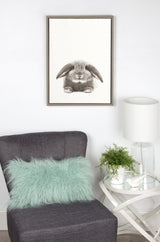 Framed gray-toned Sylvie rabbit artwork by Simon Te displayed above a gray chair with a light teal fur pillow and a round side table with a white lamp and green plant.