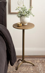 Round metal side table with a thin pedestal and three curved legs, in a warm brass finish, holding a white textured vase with green foliage and a small gold candle.