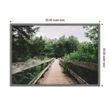 Wooden bridge pathway extending into dense green forest under an overcast sky, framed with light grey edges measuring 33 by 23 inches.