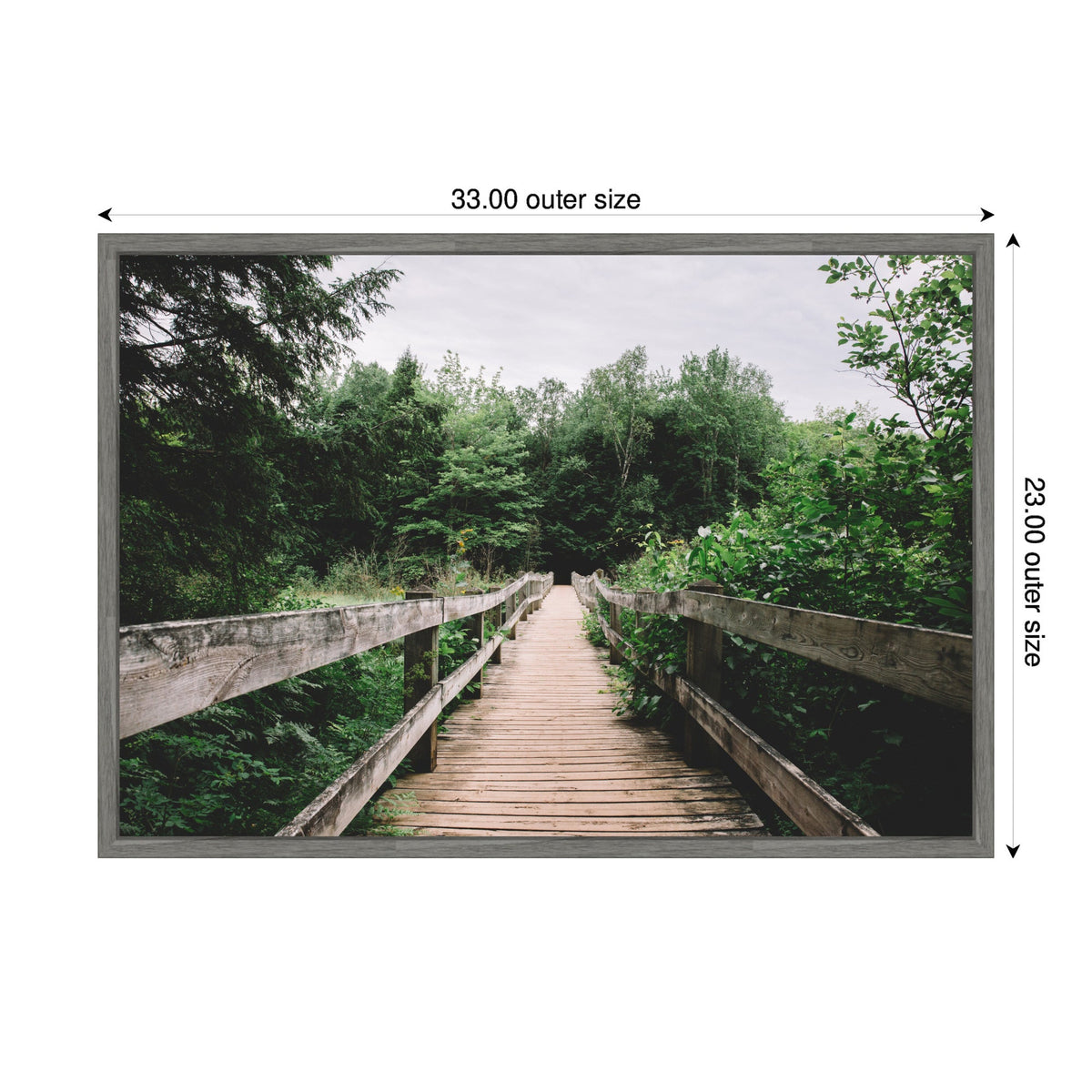 Wooden bridge pathway extending into dense green forest under an overcast sky, framed with light grey edges measuring 33 by 23 inches.