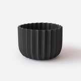 Matte black planter with vertical ridges creating a wrap-like texture and a scalloped top edge, shown against a plain white background.