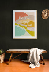 Framed abstract art print with layered pastel shapes and the text 'Be The Energy You Want To Attract' in white, hanging on a deep green wall above a rustic wooden bench with a draped white cloth.
