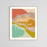 Framed art print with abstract layered shapes in terracotta, beige, mustard, pink, and turquoise, featuring the white text 'Be The Energy You Want To Attract' in a stylish serif font.