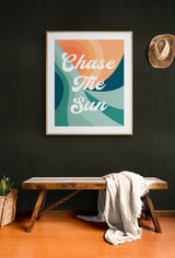 Typography art print with retro-style curved shapes in orange, teal, and green featuring white script text 'Chase The Sun' framed on a dark green wall above a wooden bench draped with a light throw.
