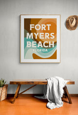 Framed modern art print with abstract shapes in warm tan, brown, and teal hues featuring text 'FORT MYERS BEACH FLORIDA' on a light gray wall above a wooden bench with a draped white cloth and a woven basket nearby.