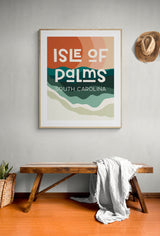 Framed modern art print with warm tones and green waves reading 'Isle of Palms South Carolina' above a wooden bench with a white throw, neutral wall and hat.
