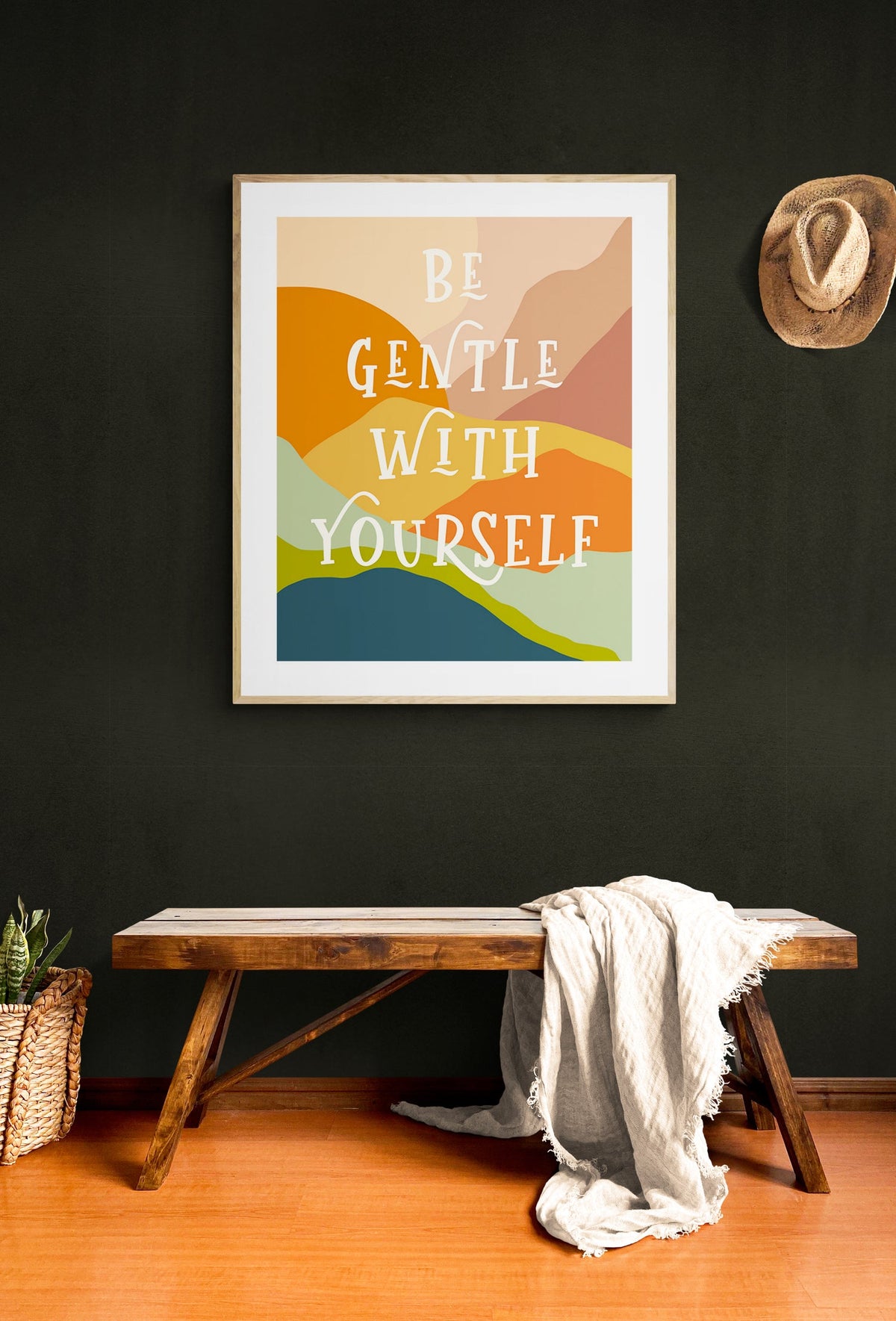 Framed art print with layered abstract shapes in orange, pink, yellow, green, and blue and white text reading 'Be Gentle With Yourself' hanging on dark wall above wooden bench with white draped cloth and straw hat on wall.