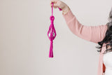 Hand holding a magenta macrame car hanging featuring a clear disco ball encased in a knotted fiber design with a tassel at the bottom.