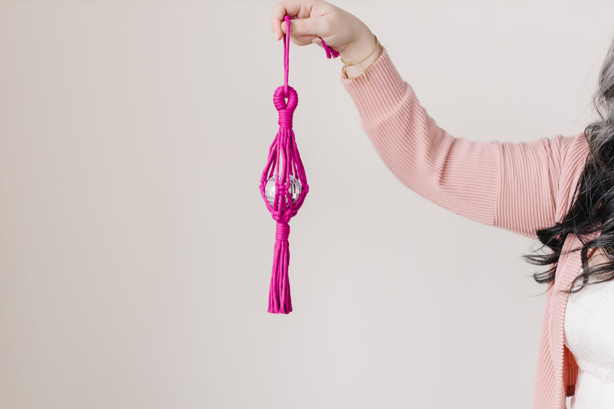 Hand holding a magenta macrame car hanging featuring a clear disco ball encased in a knotted fiber design with a tassel at the bottom.